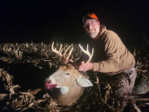 A hunter in a cornfield at night posing with a harvested white-tailed buck he is holding by the antlers.