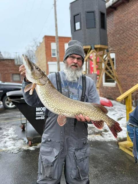 An angler holding a large northern pike in a snowy alleyway beside a brick downtown building.